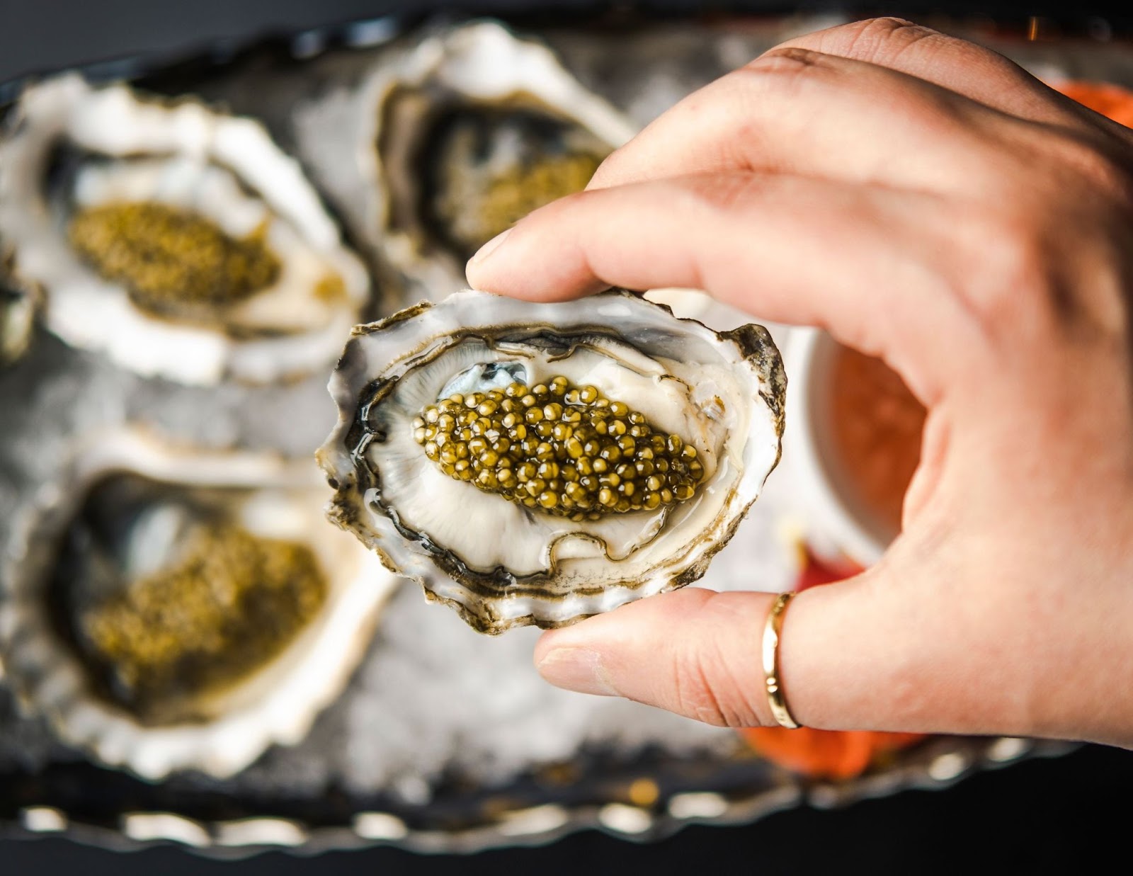 focus is on an oyster topped with caviar, held in an unidentifiable person's hand. Then there's more oysters fuzzy in the background on the plate underneath the hand. The person is wearing a gold ring on their thumb.