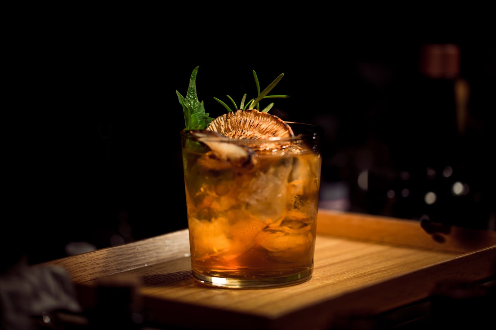 a dark, moody photo of a fancy cocktail at happy hour in kelowna, with an orange hue, dehydrated orange slice and herb garnishes