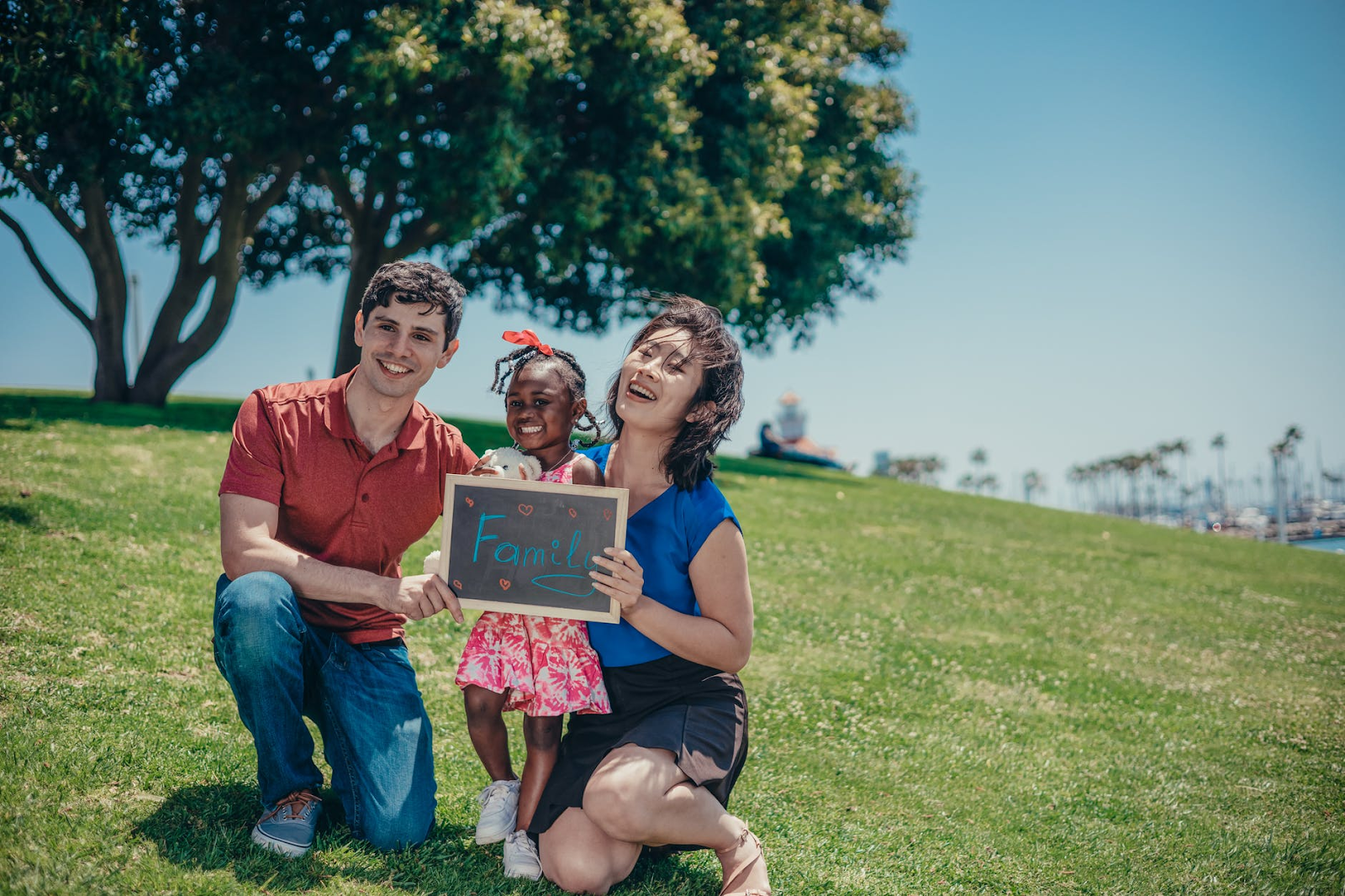 Pai, mãe e filha reunidos no parque segurando uma lousa com a palavra "family", família em inglês.