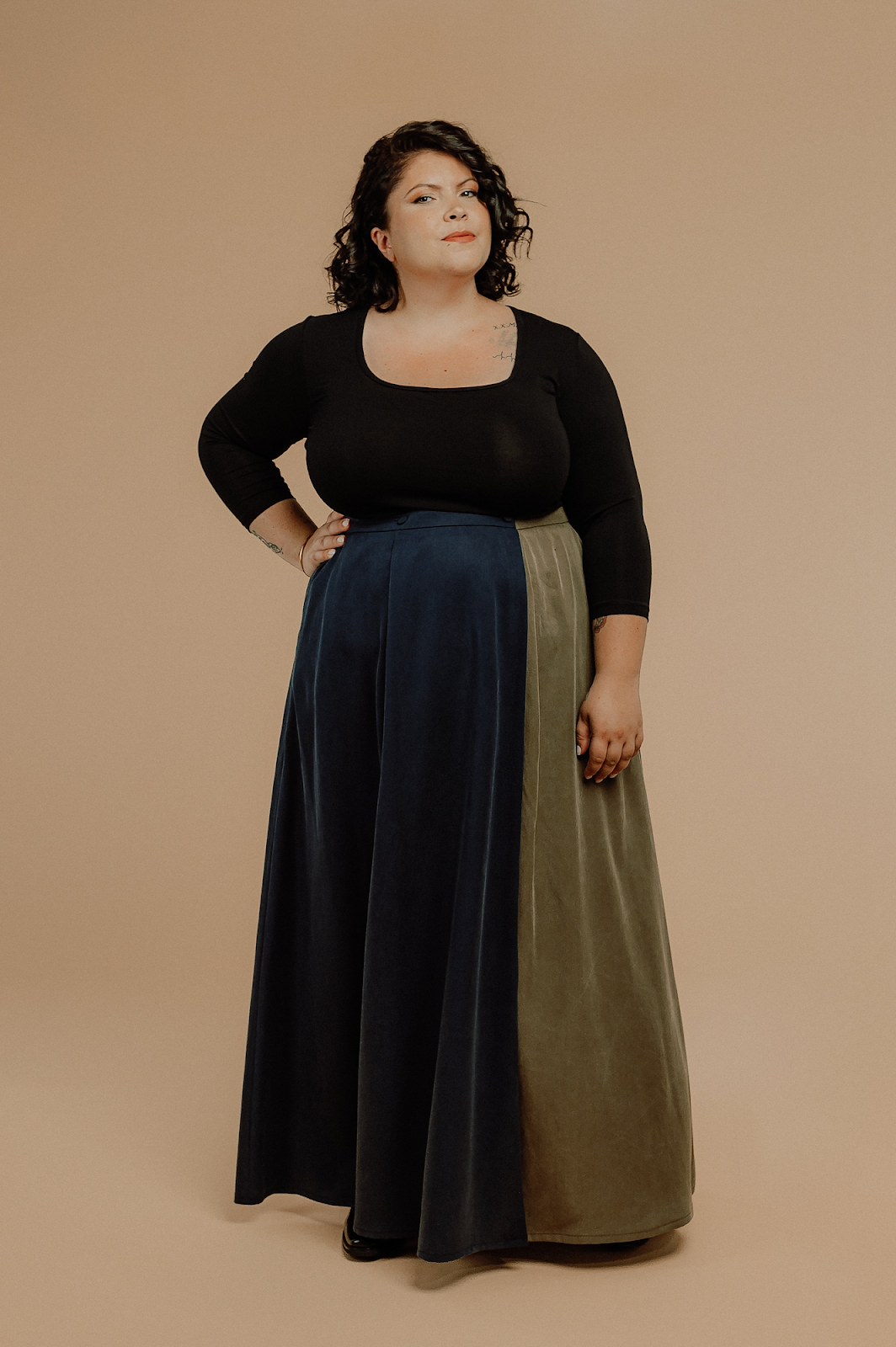 A brown person with shoulder length black hair is wearing a black 3/4 sleeve knit top with a deep squared neckline.  It is paired with a maxi skirt where the left 2/3 panel is navy and the right 1/3 panel is olive green.