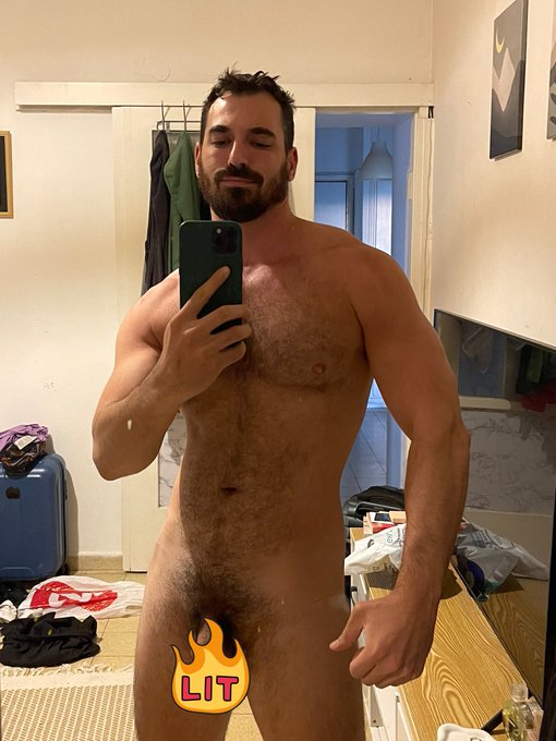 Thick Macho smiling and taking a naked iphone mirror selfie in an airbnb with a lit flame emoji covering his flaccid cock
