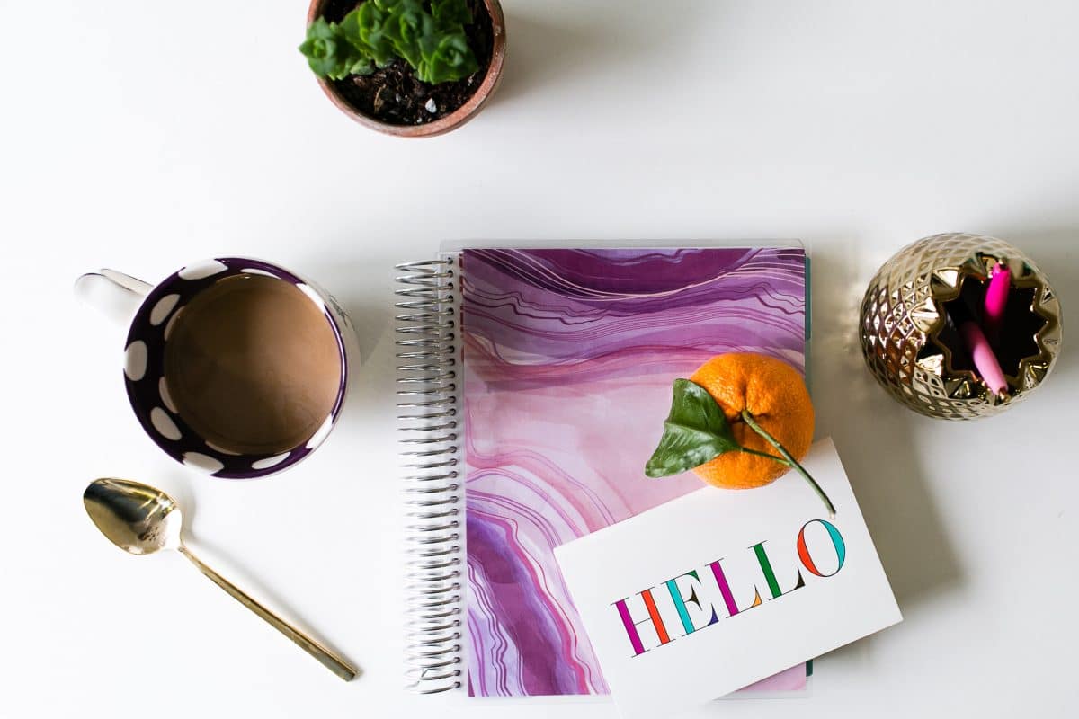 Still life image of a purple marbled planner, a cup of coffee, two plants, a greeting card and an orange on a table.