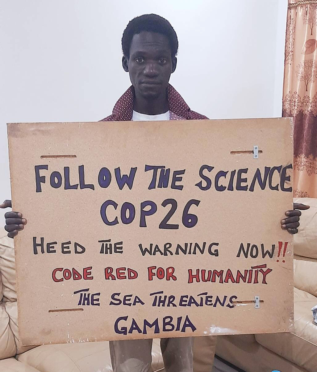 Babu stands with a large home made sign which reads 'Follow the Science COP26. Heed the warning now!! Code Red for Humanity. The sea threatens Gambia.' 