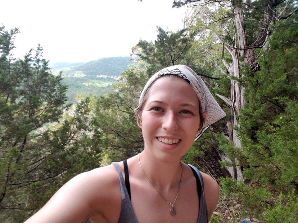 Selfie of Jessica Shainker wearing a tank top and a white bandana. In the background are trees and a scenic view of the bluffs overlooking the Buffalo River.