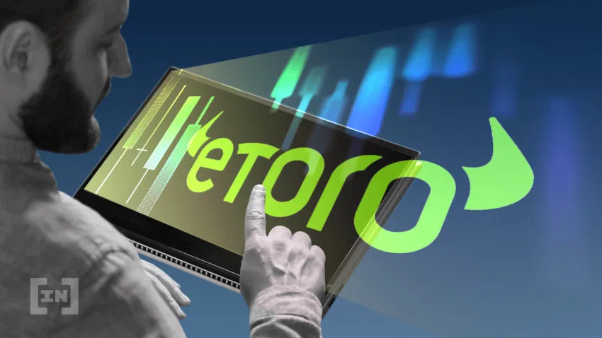 Ein Mann tippt auf einem Bildschirm, aus dem Aktienmarkt Diagramme und das Etoro Logo hervor strahlen - Ein Bild von BeInCrypto.com.