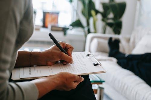 Free Unrecognizable ethnic female therapist taking notes on clipboard while filling out form during psychological appointment with anonymous client lying on blurred background Stock Photo