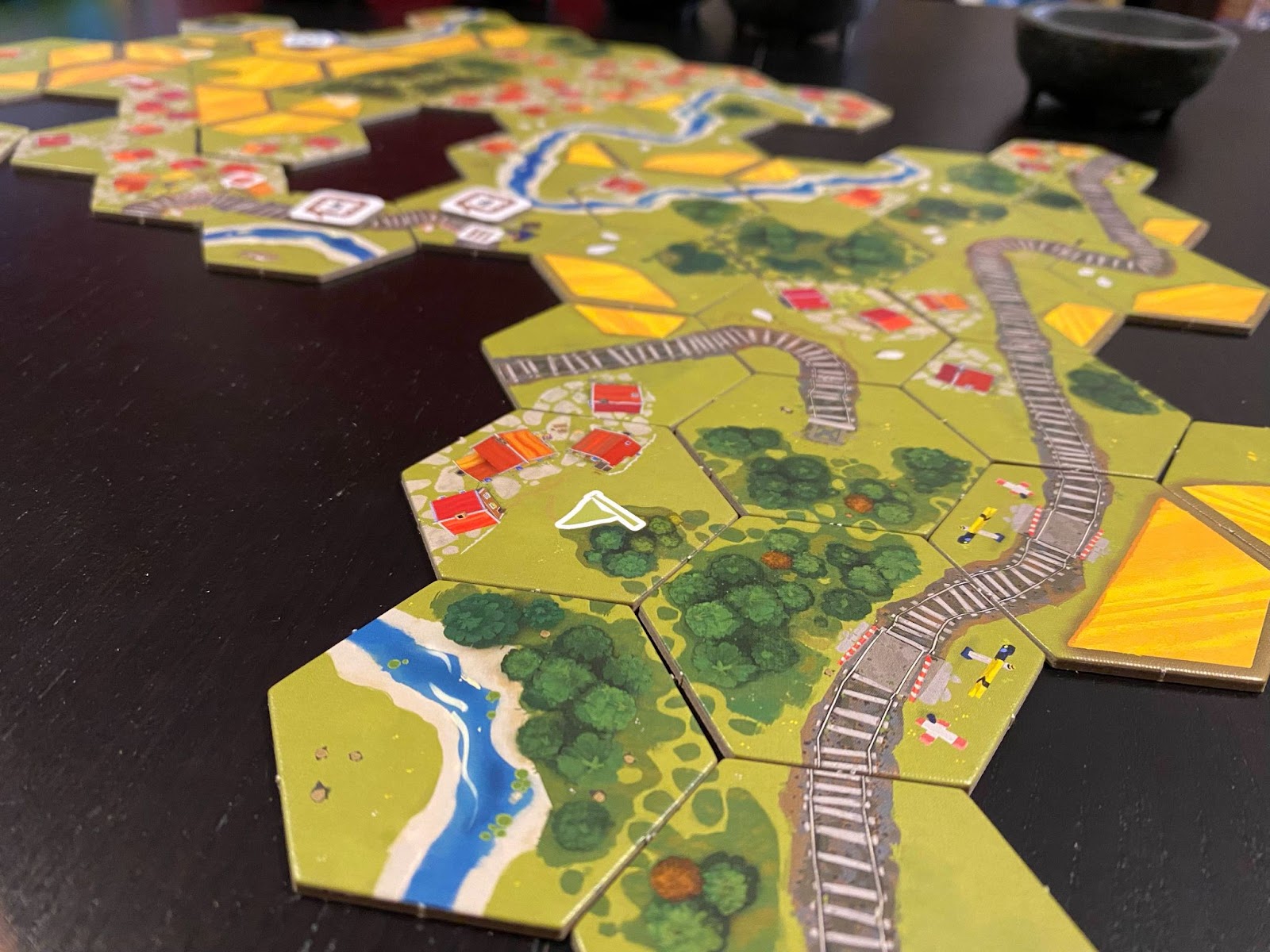 A picture of the Dorfromantik board game:  hexagonal tiles with forests, train tracks, villages and fields connected on a black table.