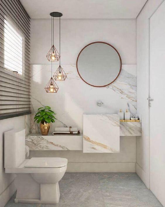 Banheiro com bancada e revestimento em meia parede da pia de porcelanato marmorizado em tom claro, piso em tons neutro, luminárias pendentes e espelho redondo.