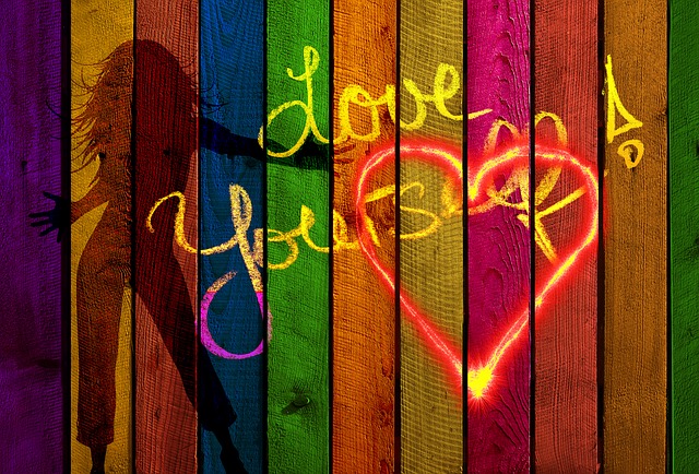 A wooden fence painted in rainbow colors, with a large outline of a heart in read and yellow, superimposed over yellow writing that says, "Love yourself!" There's a shadow on the wall of a long-haired woman with arms outstretched and one hip cocked. Loving yourself eliminates your guilt complex.