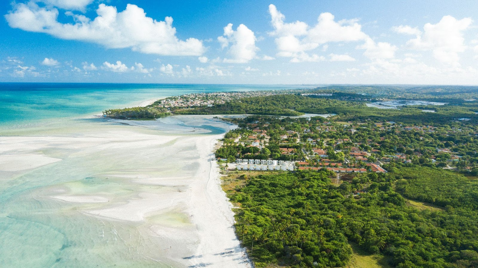 Imagem aérea da Praia do Sossego, com o mar ao lado esquerdo da foto, em frente à vegetação verde-viva ao lado direito, com algumas casas.
