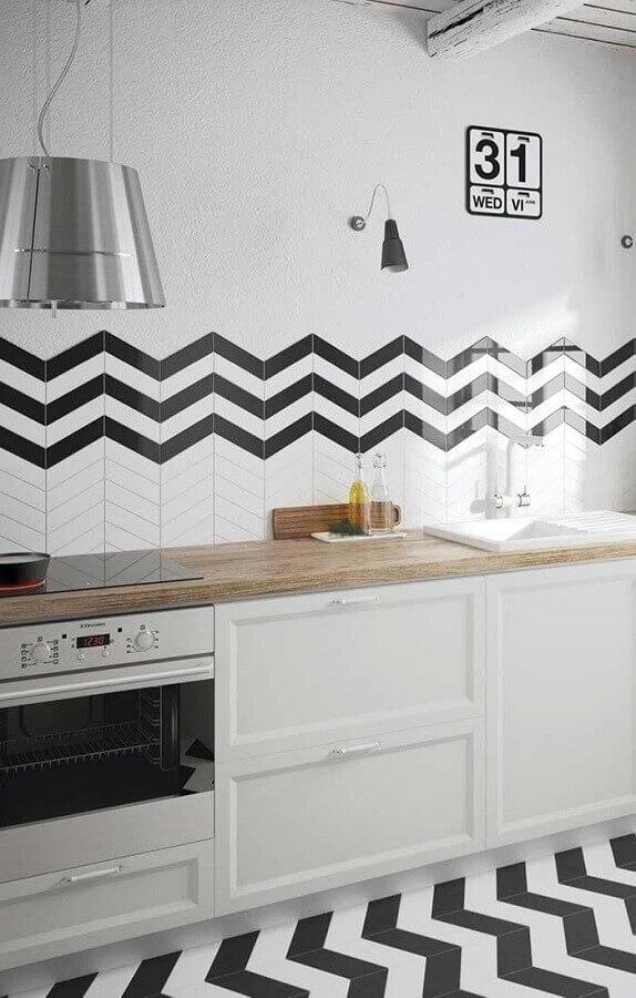 Cozinha com armários branco, bancada da pia amadeirada, luminária pendente de inox, azulejo em formato zig zag branco e preto e piso no mesmo formato branco e preto