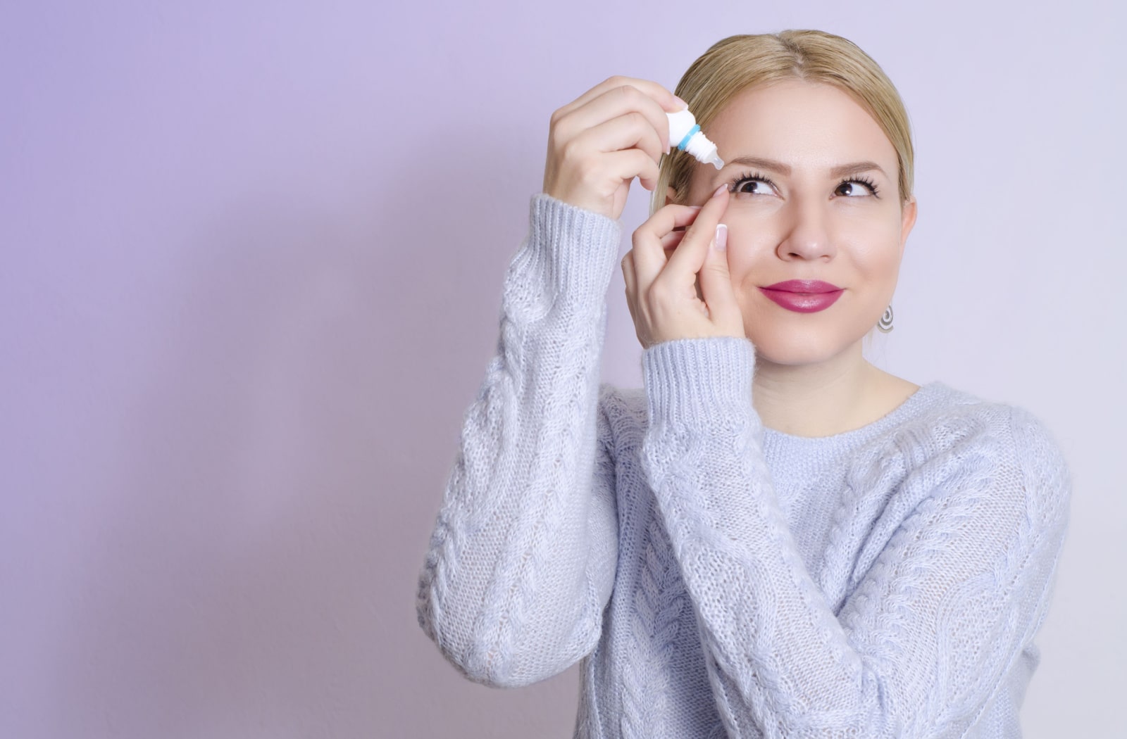 A woman wearing a lavender coloured sweater, holding her eye open to put eye drops in her eye