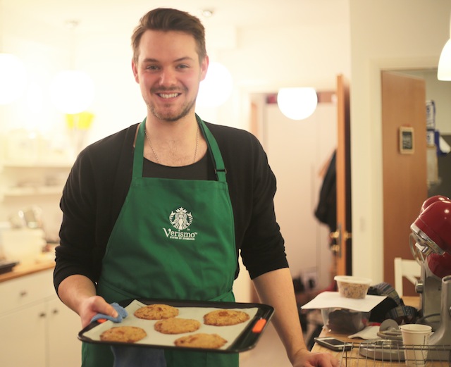 John Whaite, a baker in a black shirt and green apron smiling and holding a tray of cookies