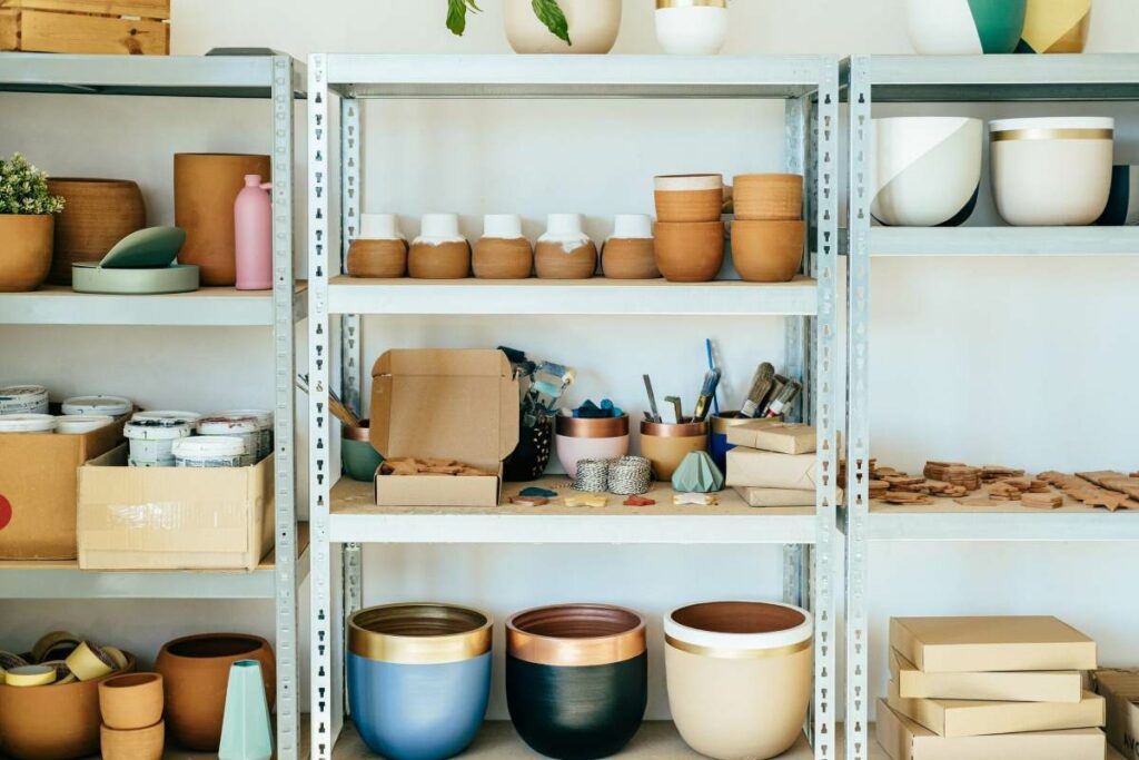 A variety of ceramics in small, medium, and large sizes, tubs of clay, and sculpting tools sit on three-tiered steel shelves.