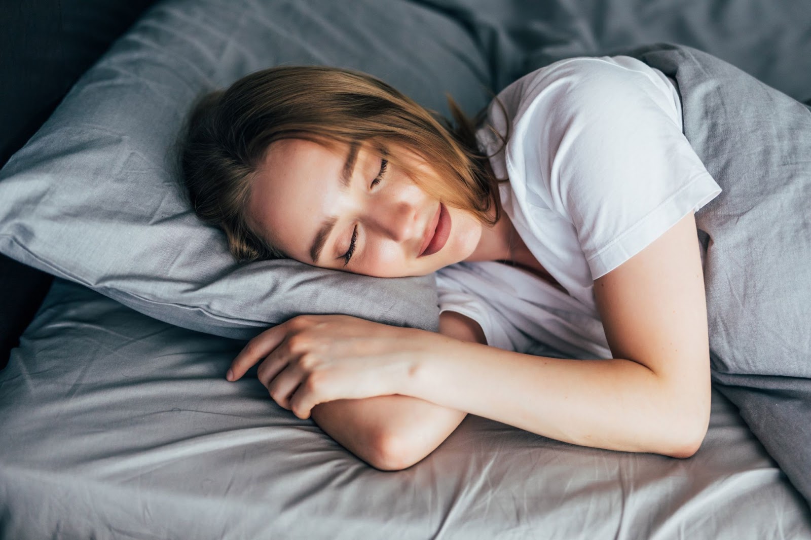 Uma mulher de pele clara e cabelos castanho claro e com uma camisa branca está de olhos fechado, deitada em uma cama com o travesseiro e o cobertor na cor cinza claro.
