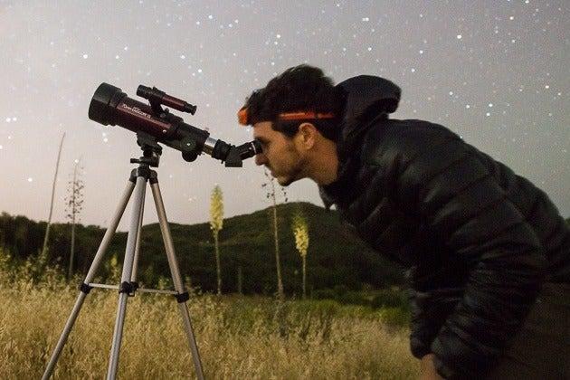 A person looking through a telescope in a field surrounded by tall grass, with fields and a starry sky in the background.