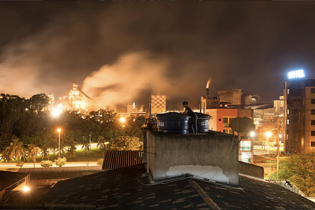A imagem mostra o telhado de uma casa. Ao fundo, é possível ver o complexo industrial da Usiminas, um céu alaranjado com uma grande nuvem de fumaça proveniente da usina da cidade.
