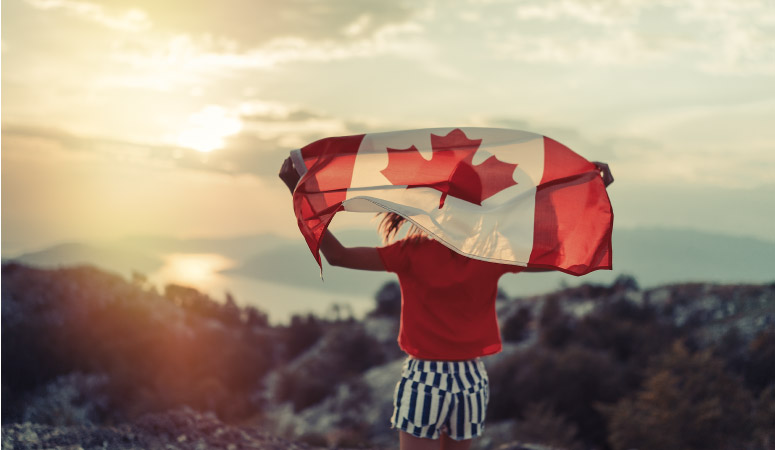 A young girl is holding the Canadian flag above her head and looking out over a stunning Canadian landscape.