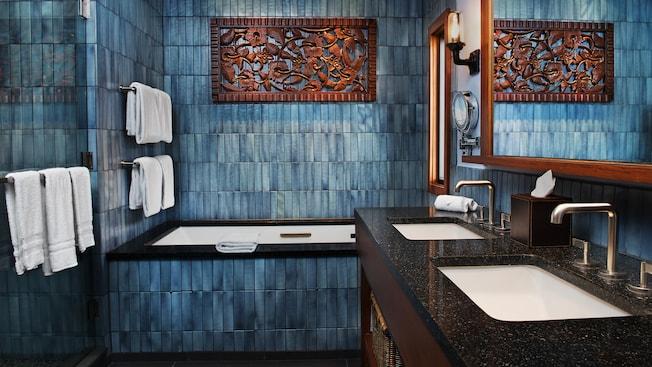 A bathroom with tile walls, a whirlpool spa bathtub, dual sinks, mirrors and Polynesian inspired art