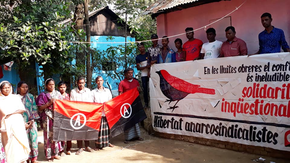 15 Bangladeshi workers holding flags and a banner of the Anarcho-Syndicalist Federation (IWA)