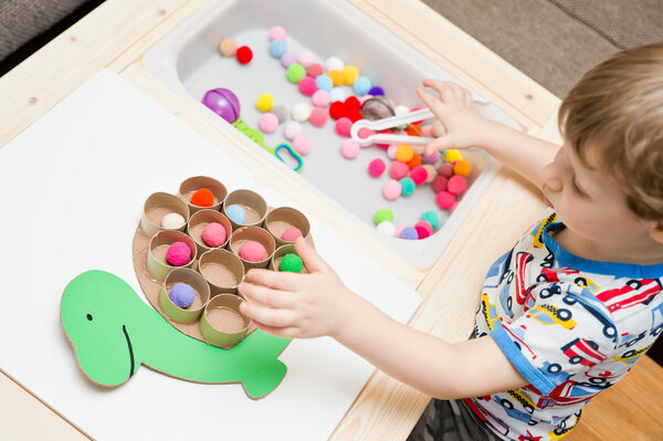A child sitting at a table with a bin of multi-colored pom poms on the right and a green animal with a brown hump that has hallow cardboard circles on it on the left. There is a pom pom in almost every hole.