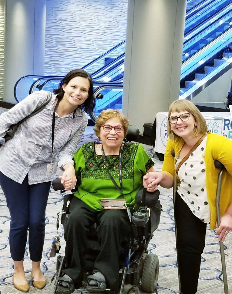 Jerri Davison and Kimberly Tissot stand and smile on both sides of Judy Heumann, who sits in her wheelchair and smiles next to them.