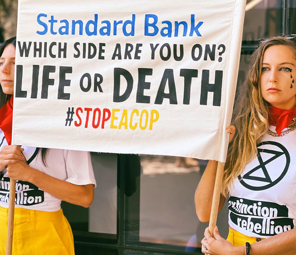 Two women wearing Extinction Rebellion t-shirts hold a banner reading 'Standard Bank - Which side are you on: Life or Death. Stop EACOP'