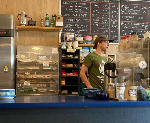 Man standing at a counter at a coffee shop with menu board behind him and baked goods beside him. Counter is blue. He's wearing a green shirt and a basebal hat