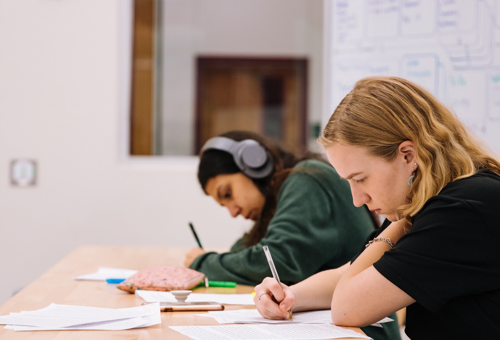 Two students sit at a table in a classroom writing on papers. A whiteboard in the background has some notes on it. One student is wearing headphones and neither are talking. Both look like they are in deep concentration.