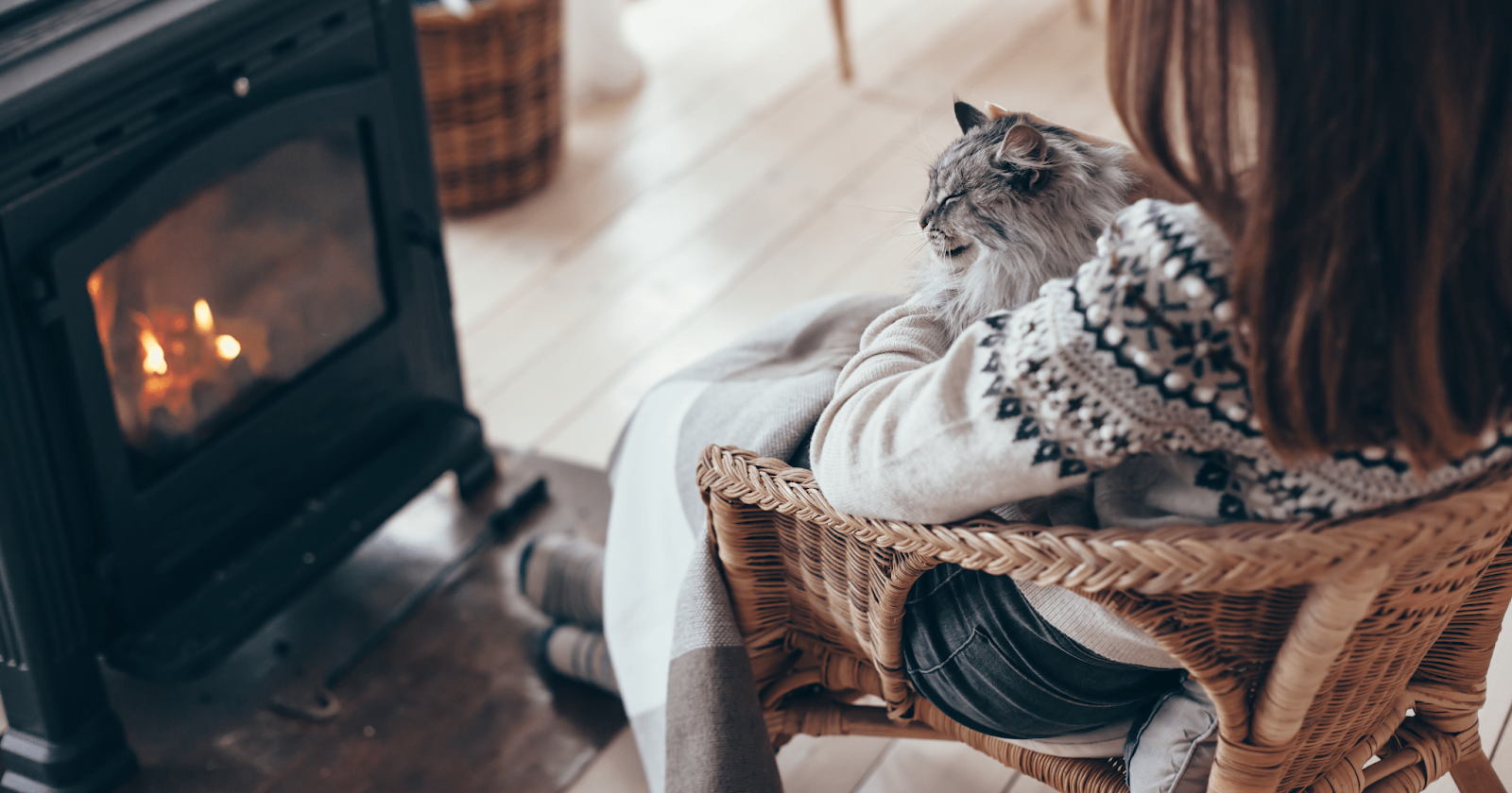 Woman sitting in whicker chair in front of fireplace with long haired cat in her lap with a blanket