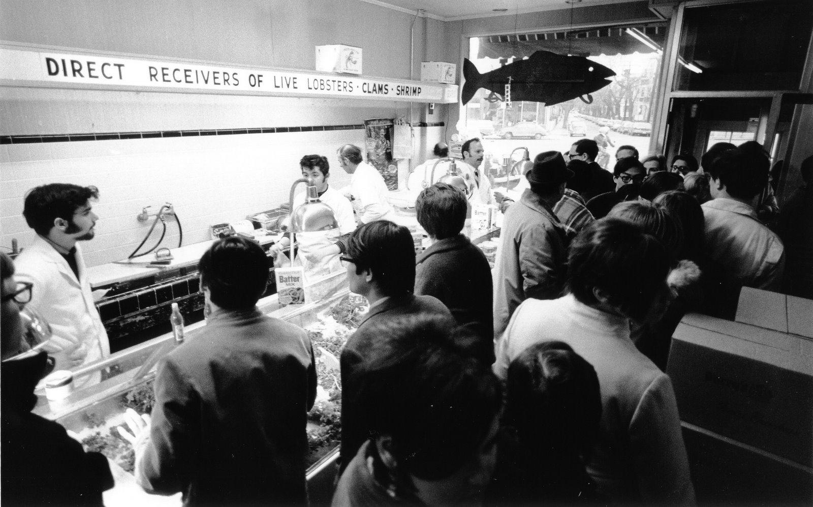 Small room filled with many people. People behind a counter are wearing white coats. Man at left has dark hair, light skin, and long sideburns. A cut out fish hangs from the ceiling.