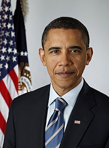 A portrait shot of Barack Obama, looking straight ahead. He has short black hair, and is wearing a dark navy blazer with a blue striped tie over a light blue collared shirt. In the background are two flags hanging from separate flagpoles: the American flag, and the flag of the Executive Office of the President.