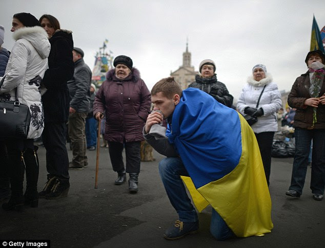 A man kneels on the ground draped in a Ukrainian flag during a funeral of an anti-government demonstrator in Independence square
