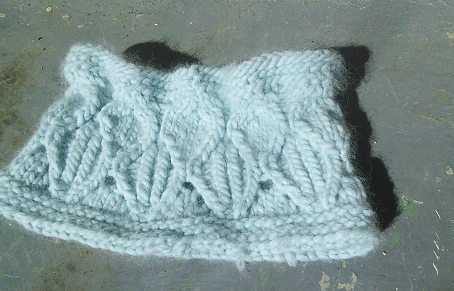 Image Description:  A lace and cable cowl worked in a bulky-weight pale blue yarn, laying flat on a concrete surface.