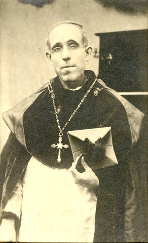 Photo noir et blanc d'un prêtre avec une grande croix pectorale, tenant sa barrette à la main.
