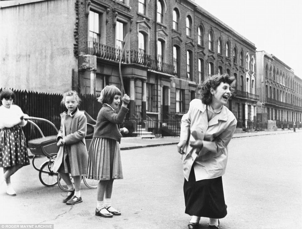 A teenager plays jump rope in the road with some younger girls. One youngster can be seen pushing an old-fashioned pram
