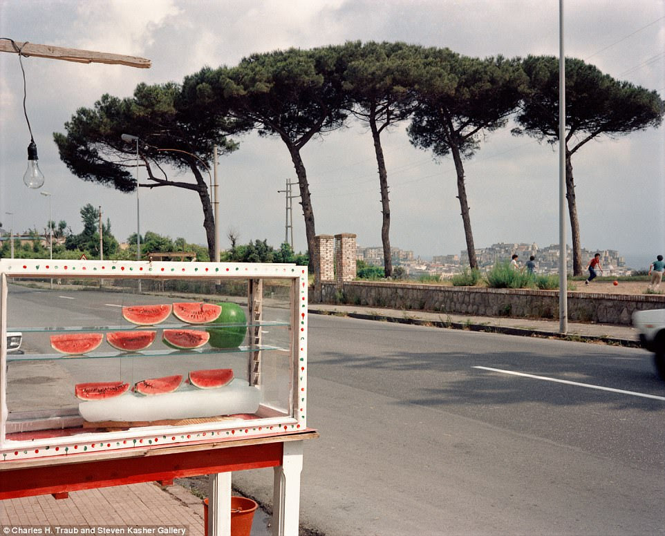 A watermelon stand on the roadside in Milan tempts passing cars. Meanwhile, a group of children play football in the background 
