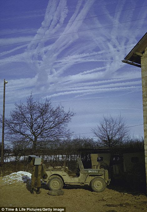Allied aircraft vapor trails in skies above US soldier unloading a jeep outside (prob.) farmhouse in the Ardennes Forest 