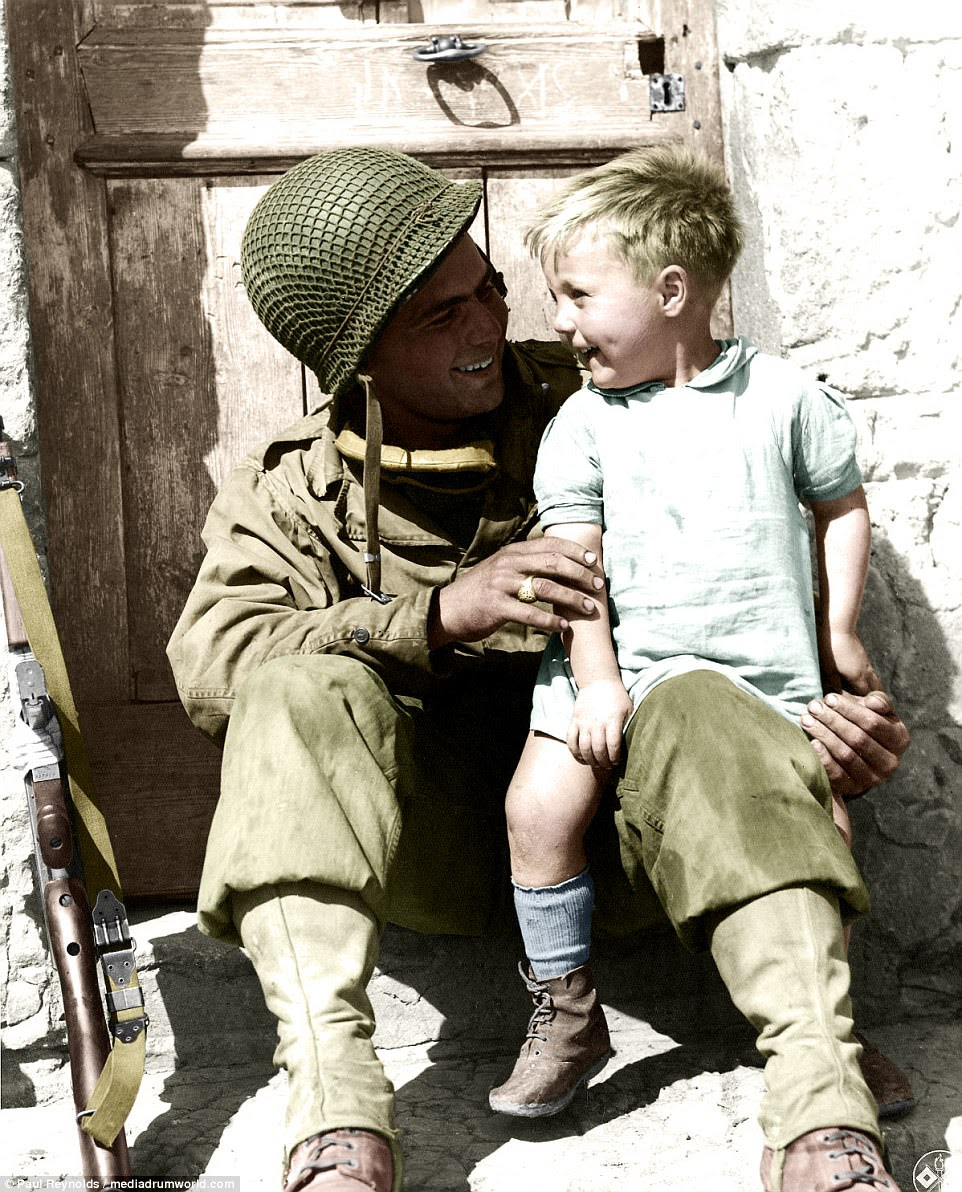 A US soldier laughs with a young boy as he puts his gun to one side and sits the youngster on his knee on a front doorstep in a French village in July 1944, in one of a series of striking colourised images of children caught up in the two world wars