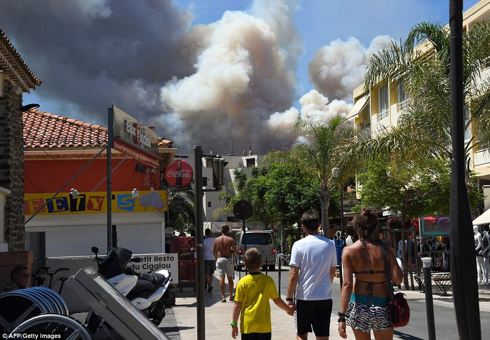 People walk through the streets of Bormes-les-Mimosas, southeastern France, as smoke rises from a fire burning a forest near the seaside town