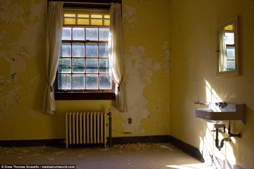 Yellow paint flakes off the walls in a room with a sink in the psychiatric hospital. The curtains can be seen drawn above a radiator