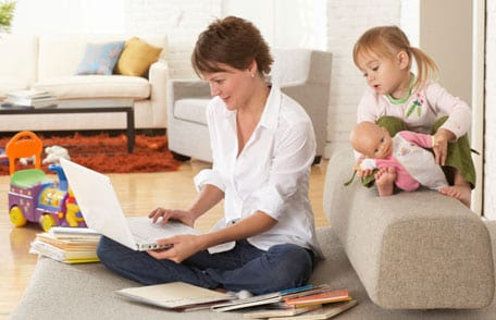 Mother using laptop while daughter plays with toys behind her