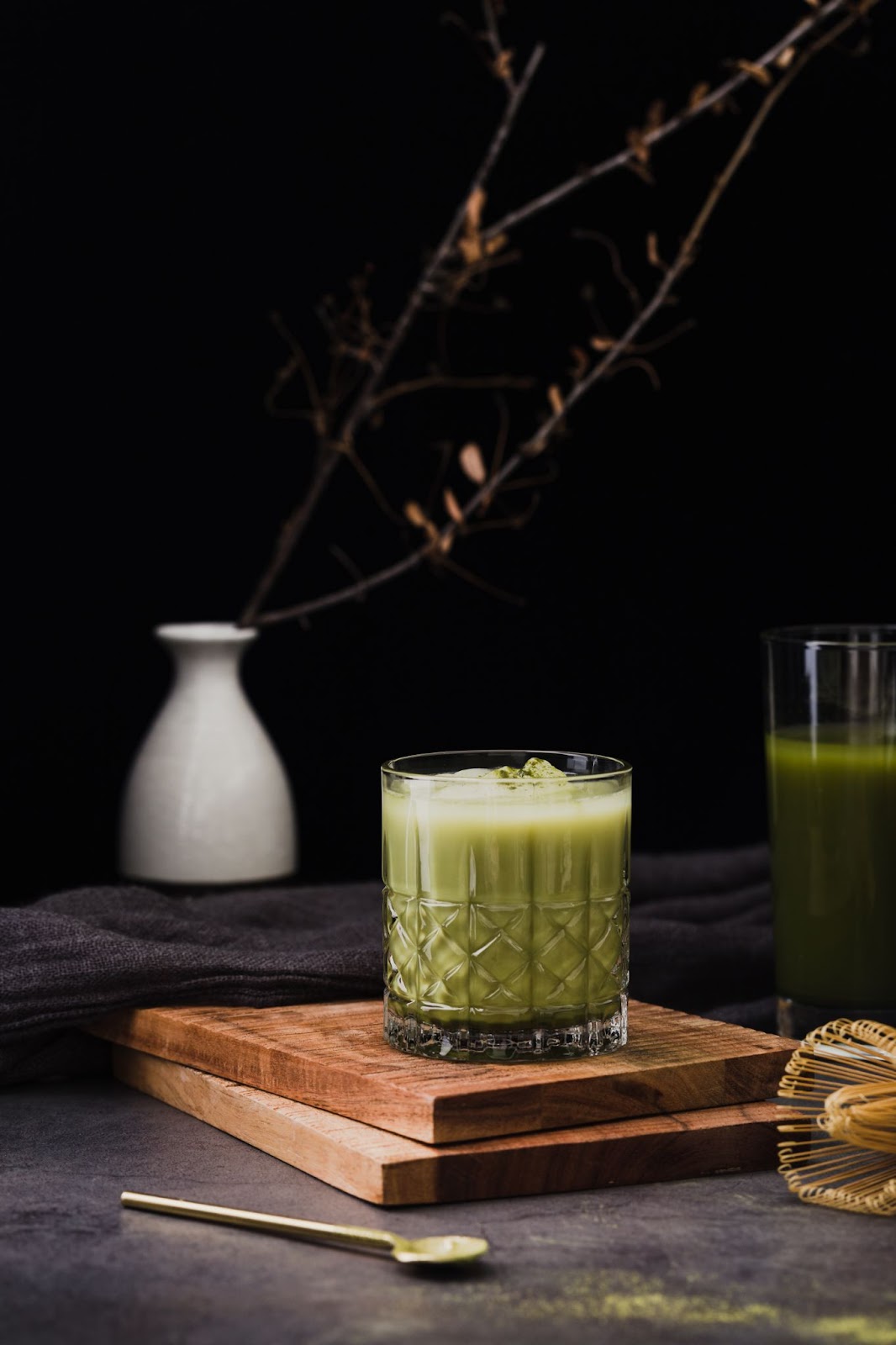 a imagem mostra um chá verde gelado com cubos de gelo dentro, em cima de duas tábuas de madeira. Ao redor há outro copo com chá verde e uma colher de chá.