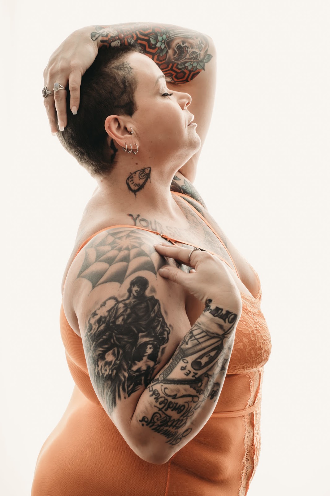 Plus sized woman with shaved head and tattoos stands in front of a high key background. She's wearing an orange bodysuit with lace. Photo by Embodied Art Boudoir. Classic boudoir photography, classic photography, classic boudoir, classy boudoir, classy photography, colorado boudoir, denver boudoir, boulder boudoir, colorado springs boudoir, boudoir ideas, boudoir poses, boudoir inspiration, photography inspiration, boudoir session, boudoir photos