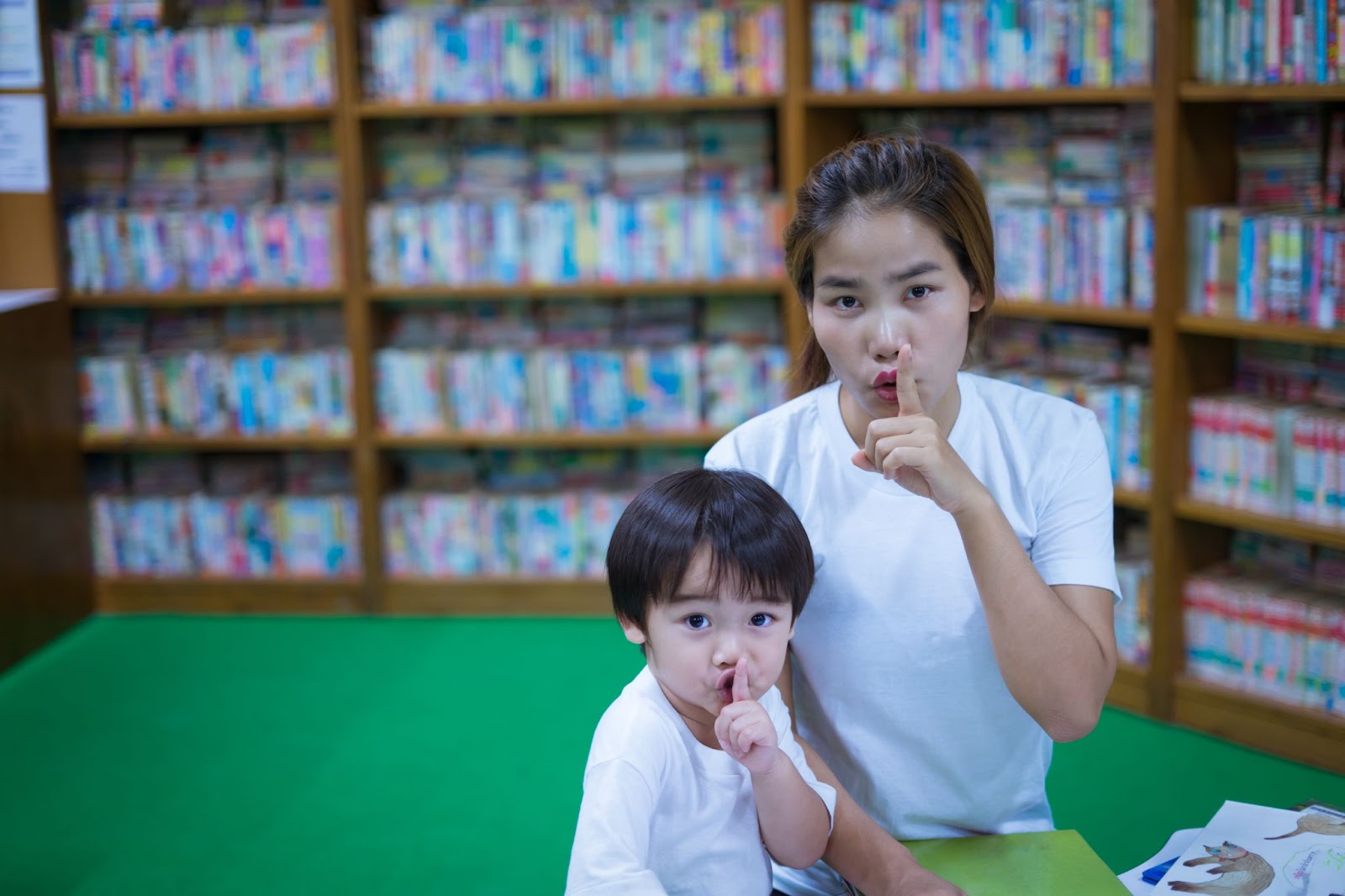 Asian teacher with little boy reading book and holding finger on lips making a silent gesture together in library.