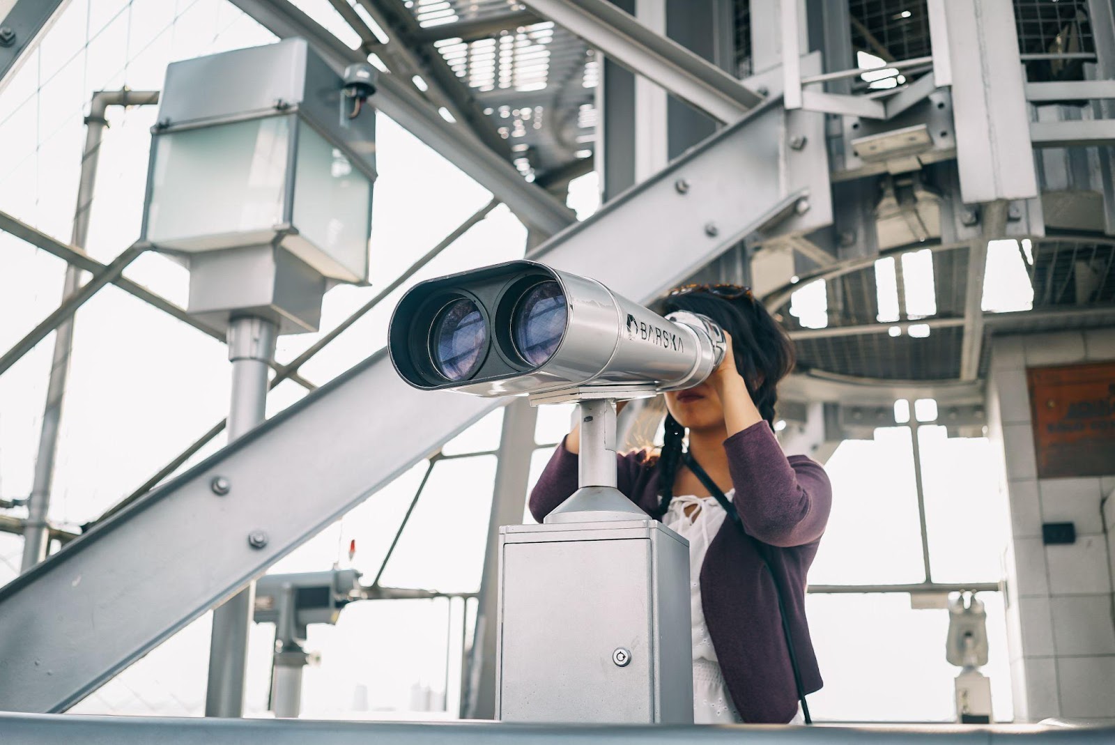 A person gazes into a high tech telescope standing in front of what appears to be a technical or engineering related object made from metal.