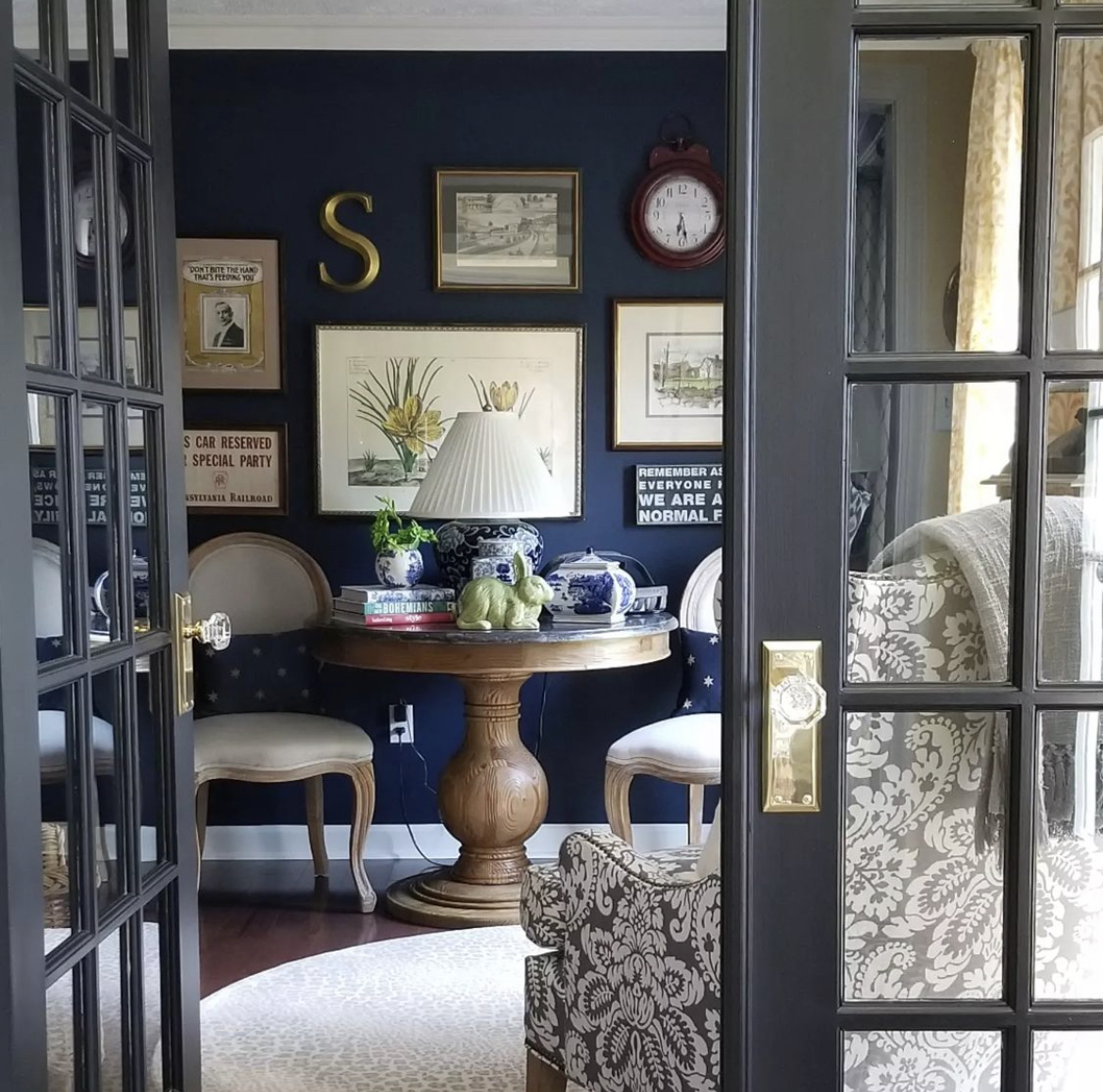 A stunning study with deep rich blue painted wall features a gallery wall full of prints and decorative items, a round table decorated with blue and white porcelain pieces and 2 side chairs 