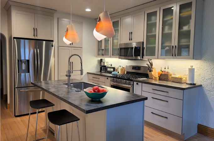 A newly remodeled kitchen with a combination of traditional cabinets and frosted window cabinets, all painted in a neutral gray. There is also a two-seater island with a built-in sink, and new chrome appliances throughout the kitchen. 