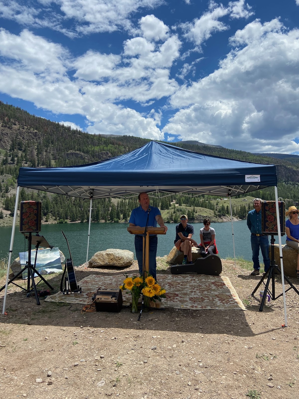 Governor Polis at a podium under a shade tent by a lake.