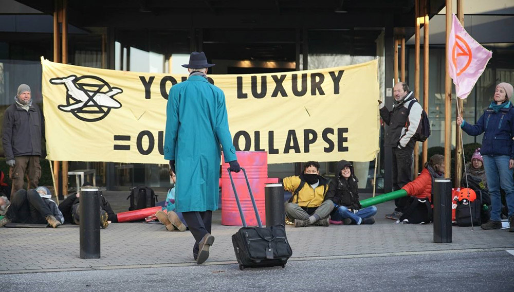 Traveller in blue raincoat and pulling a wheelie suitcase walks towards rebels holding a banner reading 'Your Luxury = Our Collapse'